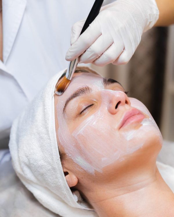 Beautician with a brush applies a white moisturizing mask to the face of a young girl client in a spa beauty salon